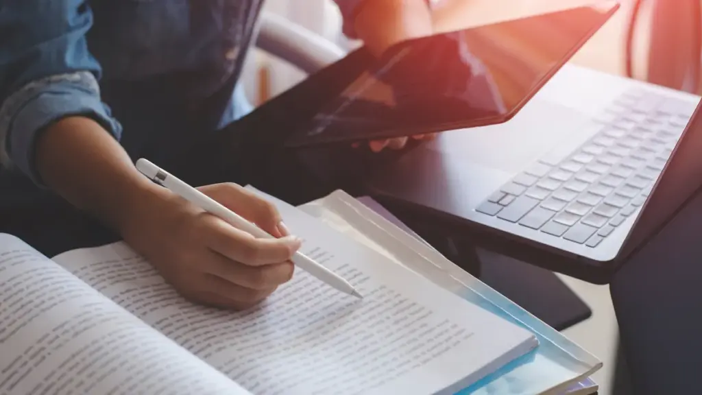 Persona leyendo un libro abierto con lápiz digital, junto a una tablet y un portátil sobre la mesa